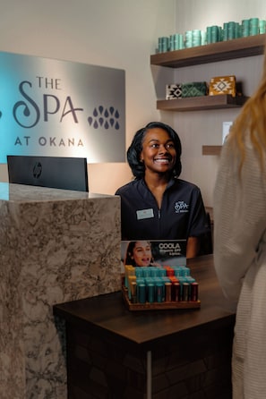 A reception desk with a sign for 'The Spa at Okana' and a display of 'COOLA Organic SPF Lipux.' - OKANA Resort & Indoor Waterpark (Oklahoma City)