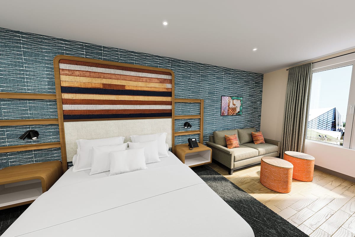 Ein modernes Hotelzimmer mit einem großen Bett, einer Couch, einem kleinen Hocker und Blick auf die Stadt durch das Fenster.