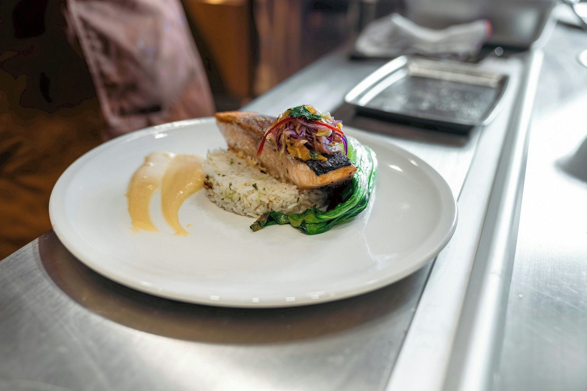 A white plate with a fish fillet, rice, and a green vegetable on a stainless steel table.
