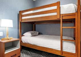 A bunk bed with a wooden frame, a bedside table with a lamp, and a gray wall.