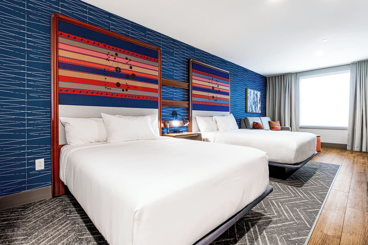 Ein Hotelzimmer mit zwei Betten, einem großen Fenster mit Vorhängen und einer Wand mit blauem Backsteinmuster.