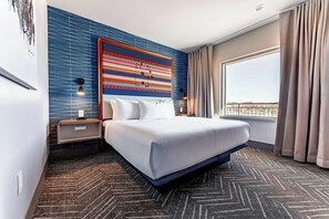 A hotel room with a large bed, a nightstand, a wall art piece, a window with curtains, and a patterned carpet. - OKANA Resort & Indoor Waterpark (Oklahoma City)