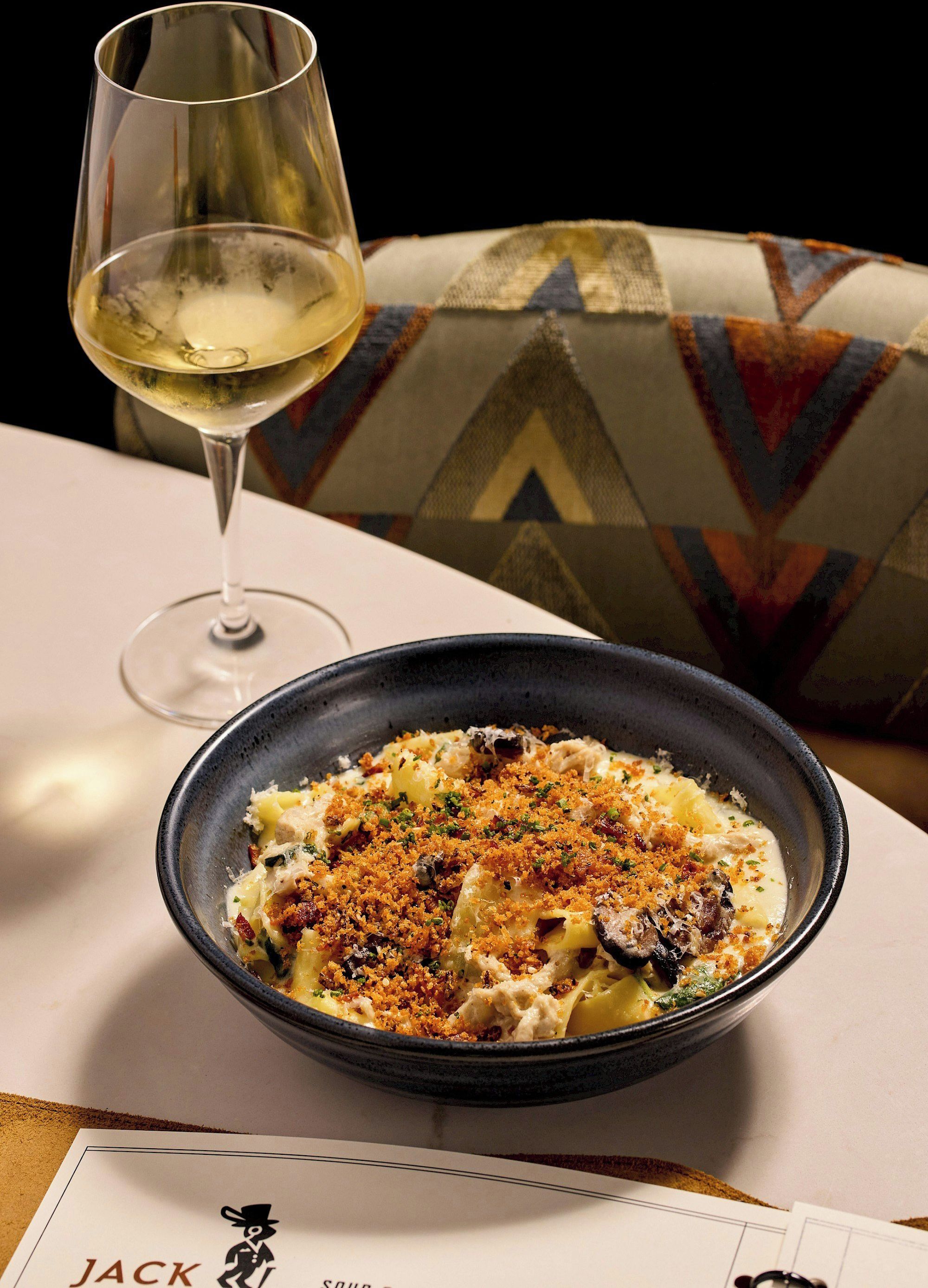 Eine Schüssel Pasta mit Streuseln, ein Glas Weißwein und eine Speisekarte auf einem Tisch mit einem gemusterten Kissen im Hintergrund.