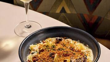 Eine Schüssel Pasta mit Streuseln, ein Glas Weißwein und eine Speisekarte auf einem Tisch mit einem gemusterten Kissen im Hintergrund.