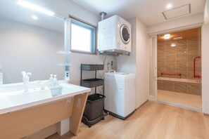 A modern bathroom with a built-in washing machine, a shower with a red handrail, and a sink with a mirror. - Naha Shintoshin Hotel (Naha)