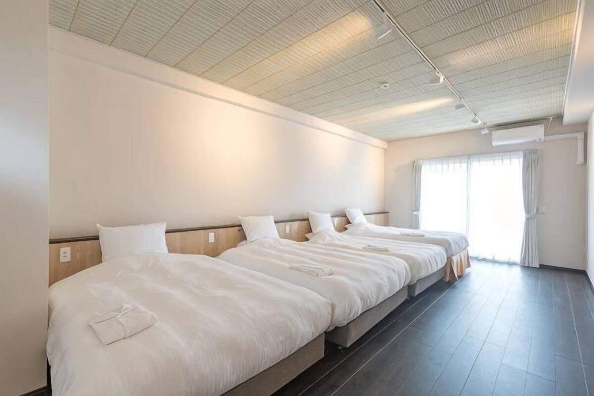 Ein Hotelzimmer mit einer Reihe von Betten, Fliesenboden und einem großen Fenster mit Vorhängen.
