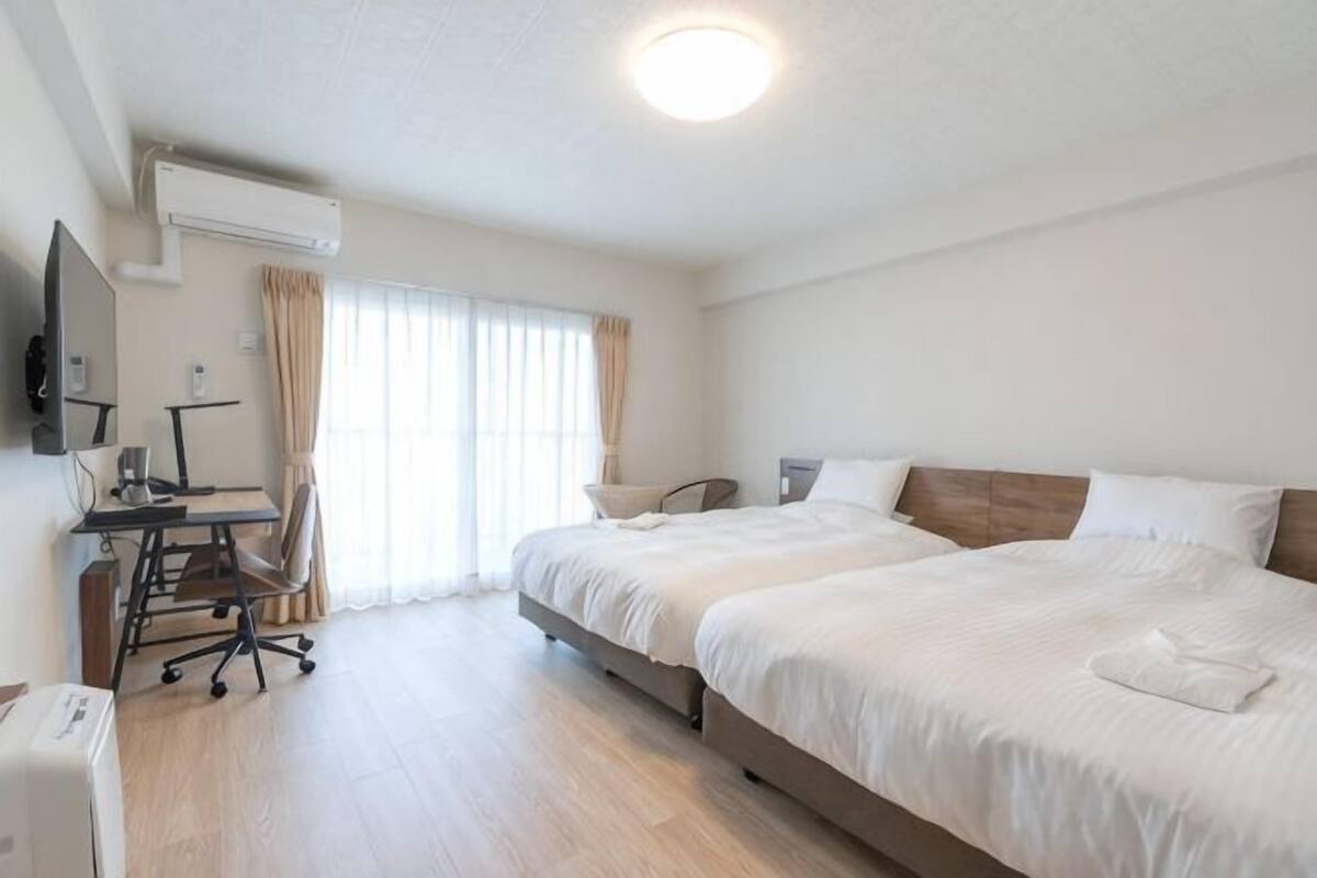 Ein Hotelzimmer mit zwei Betten, einem Schreibtisch mit Stuhl, einem Flachbildfernseher und einer Klimaanlage.