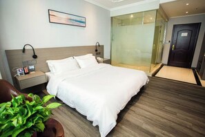 Habitación de hotel con cama, mesita de noche, planta en maceta y cabina de ducha de vidrio, además de una puerta.