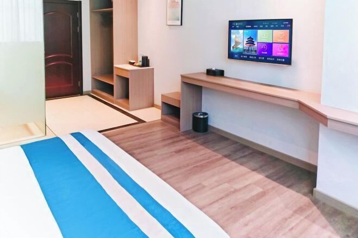 A hotel room with a bed, a TV mounted on the wall, a wooden floor, and a small desk area.