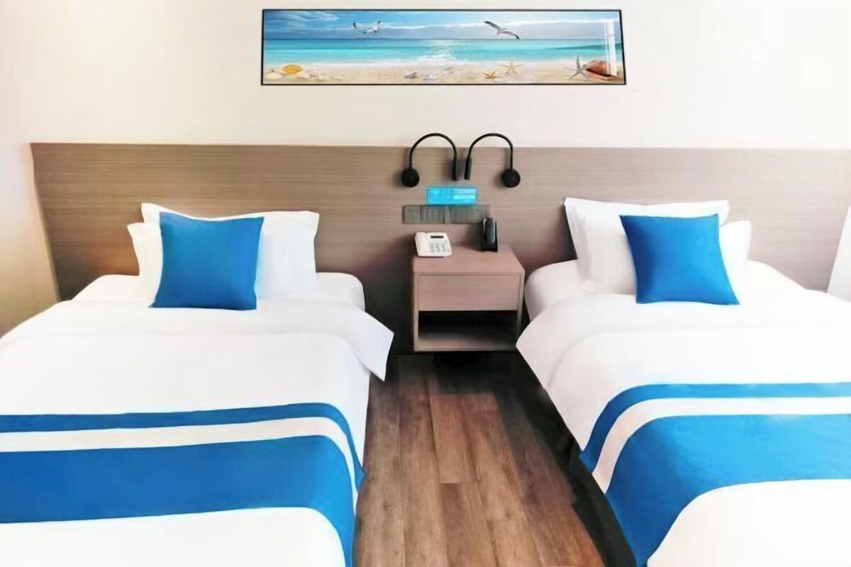 A hotel room with two beds, each with blue and white bedding, a wooden headboard, a nightstand with a phone and headphones, and a beach-themed painting on the wall.