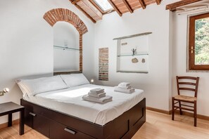 A bedroom with a bed, a wooden chair, a small table, a skylight, and a brick wall.