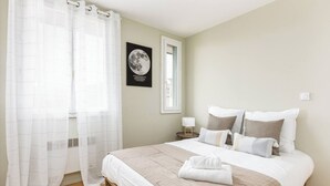 A neatly made bed with a beige and white color scheme, a framed picture of the moon on the wall, and a window with a view of the outside.