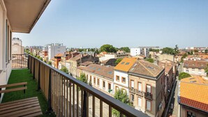 A balcony with a view of a cityscape, featuring a mix of historic and modern buildings, a clear sky, and a green lawn area.