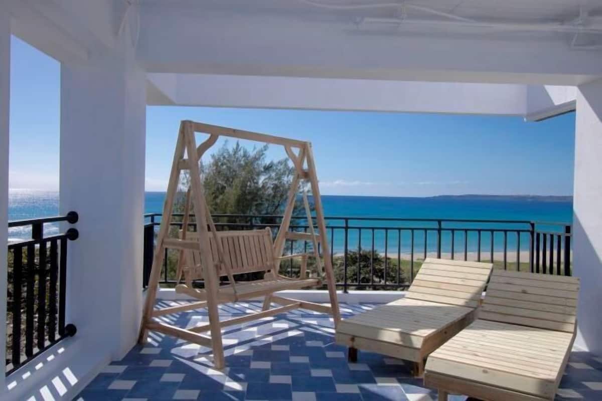 A balcony with a swing chair, two lounge chairs, and a view of the sea.