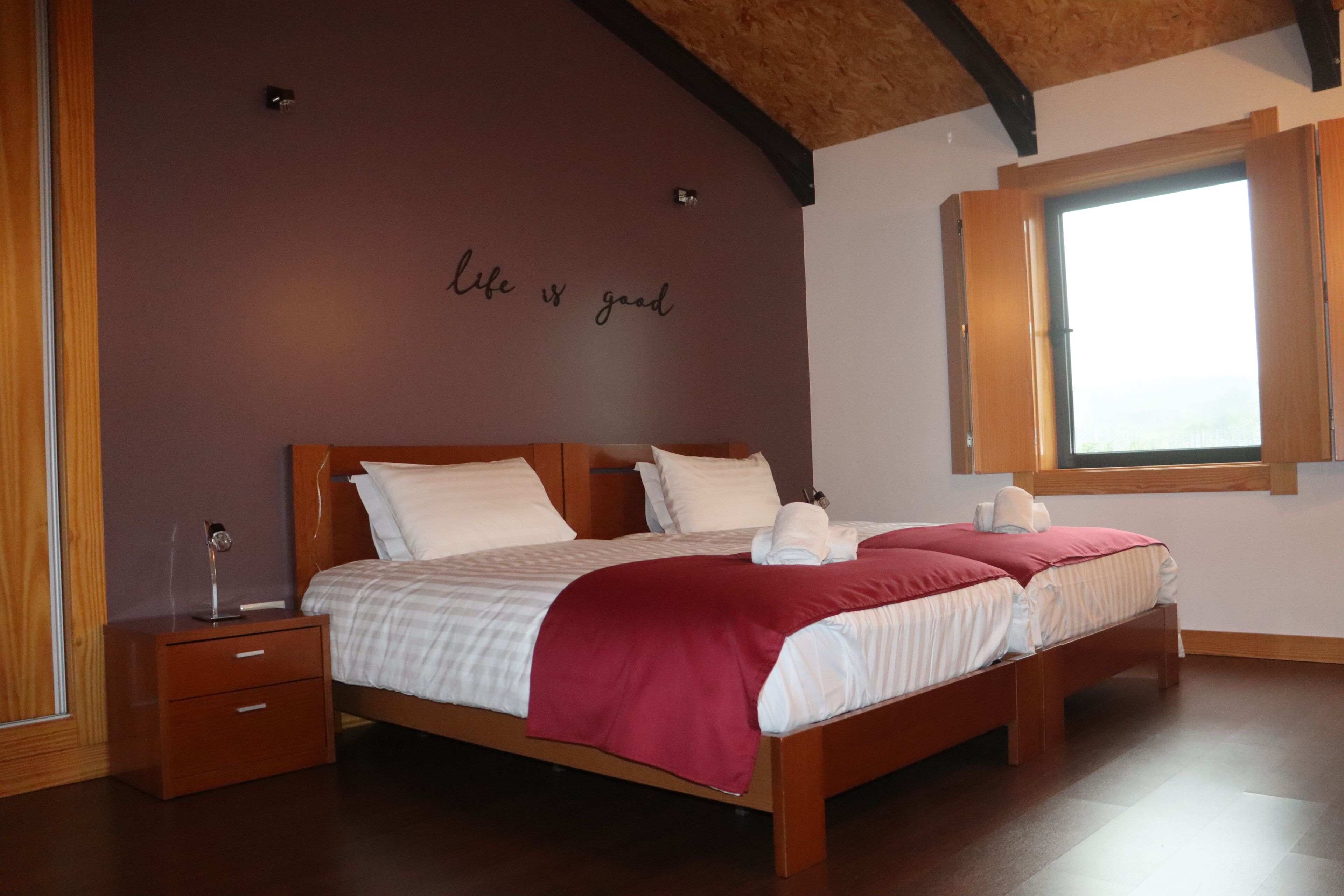 Quarto com cama de madeira, mesa de cabeceira, janela com persianas de madeira e parede com frase motivacional.