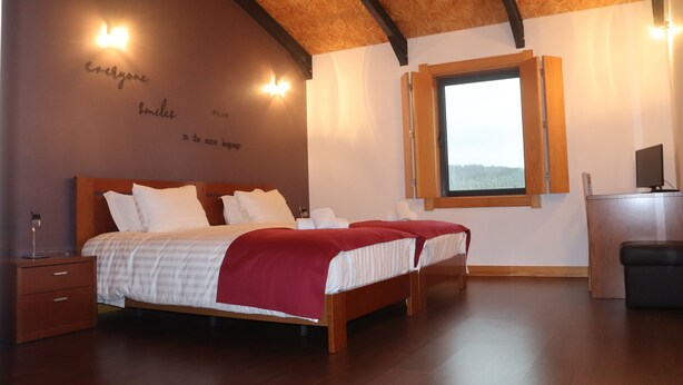 Une chambre avec un plafond en bois, un grand lit avec une literie blanche et rouge, une table de chevet, un bureau et une fenêtre donnant sur un beau paysage.