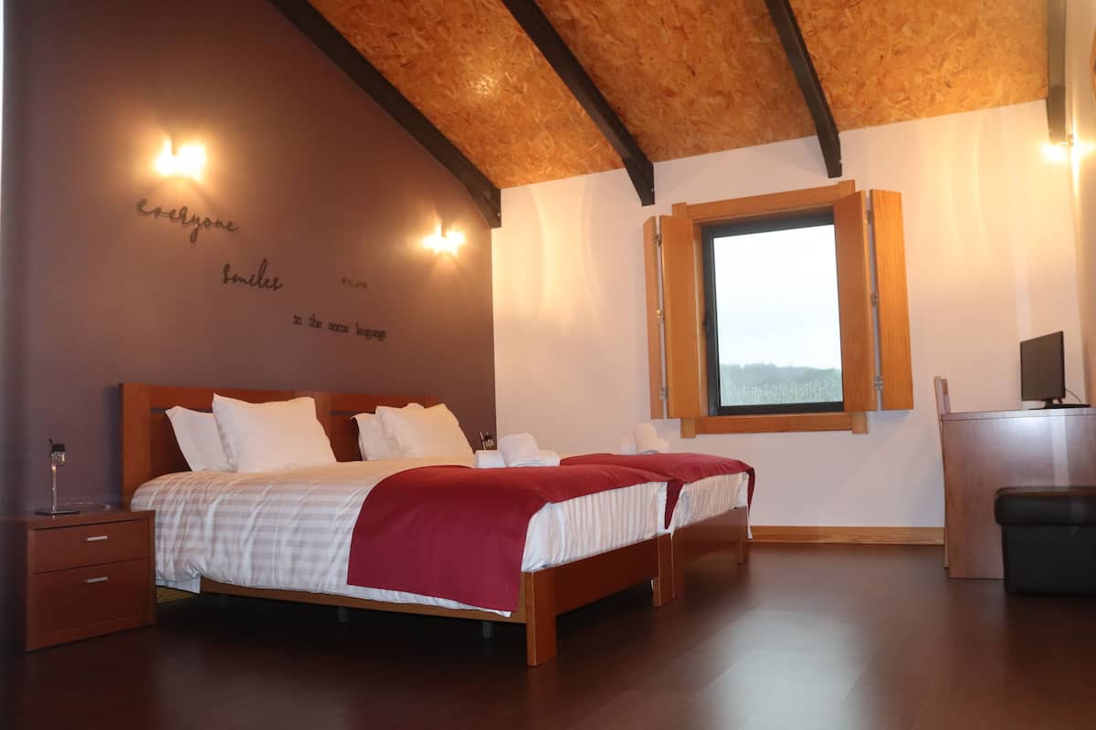 A bedroom with a wooden ceiling, a large bed with white and red bedding, a nightstand, a desk, and a window with a view.