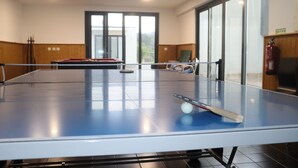Une table de ping-pong se trouve dans une pièce dont le sol est en bois, où l’on aperçoit également un extincteur et une porte donnant sur l’extérieur.