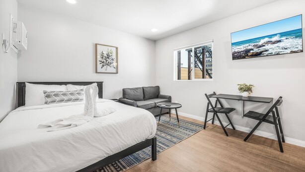 Une chambre d’hôtel moderne avec un lit, un canapé gris, une petite table et un bureau avec deux chaises. Il y a une fenêtre donnant sur la mer et un tableau encadré au mur.