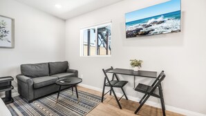 Un salon moderne avec un canapé gris, une petite table ronde et un coin repas comprenant une table et des chaises aux couleurs sombres. Une grande fenêtre offre une vue sur l’océan, et un tableau encadré orne le mur.