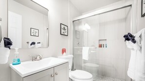 Une salle de bain moderne avec un lavabo, des toilettes et une douche, tous de couleur blanche. Des serviettes sont accrochées au mur et un tableau encadré est placé au-dessus du lavabo.