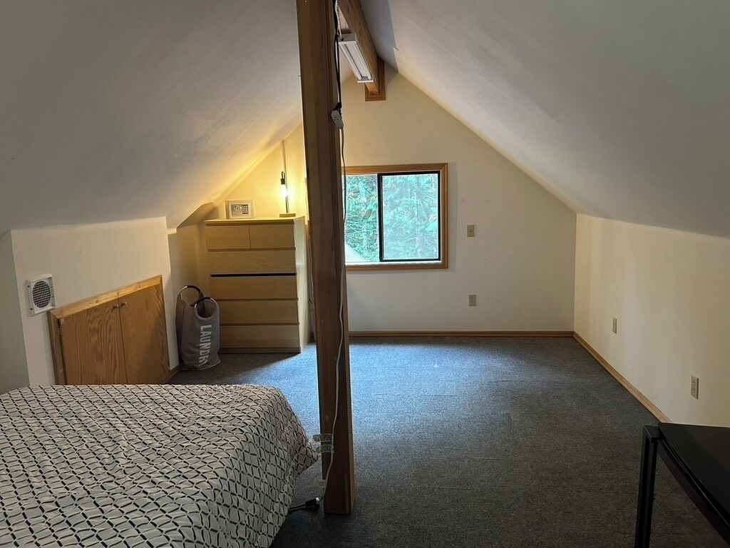 Un dormitorio con techo inclinado, una ventana, una cama, un aparador de madera y una bolsa en el suelo.