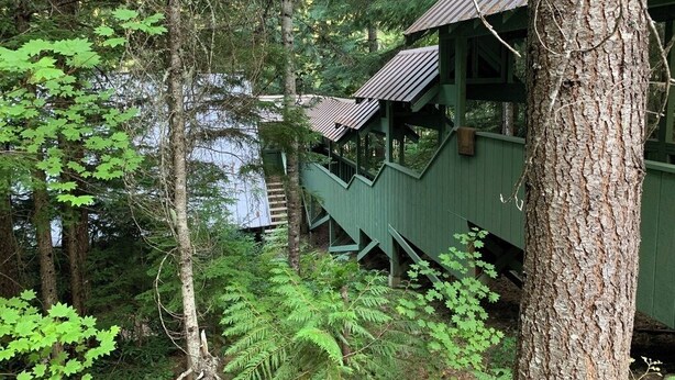Eine Holzhütte mit einer Treppe, die zu einer Plattform führt, umgeben von dichtem Wald und einem Gewässer.
