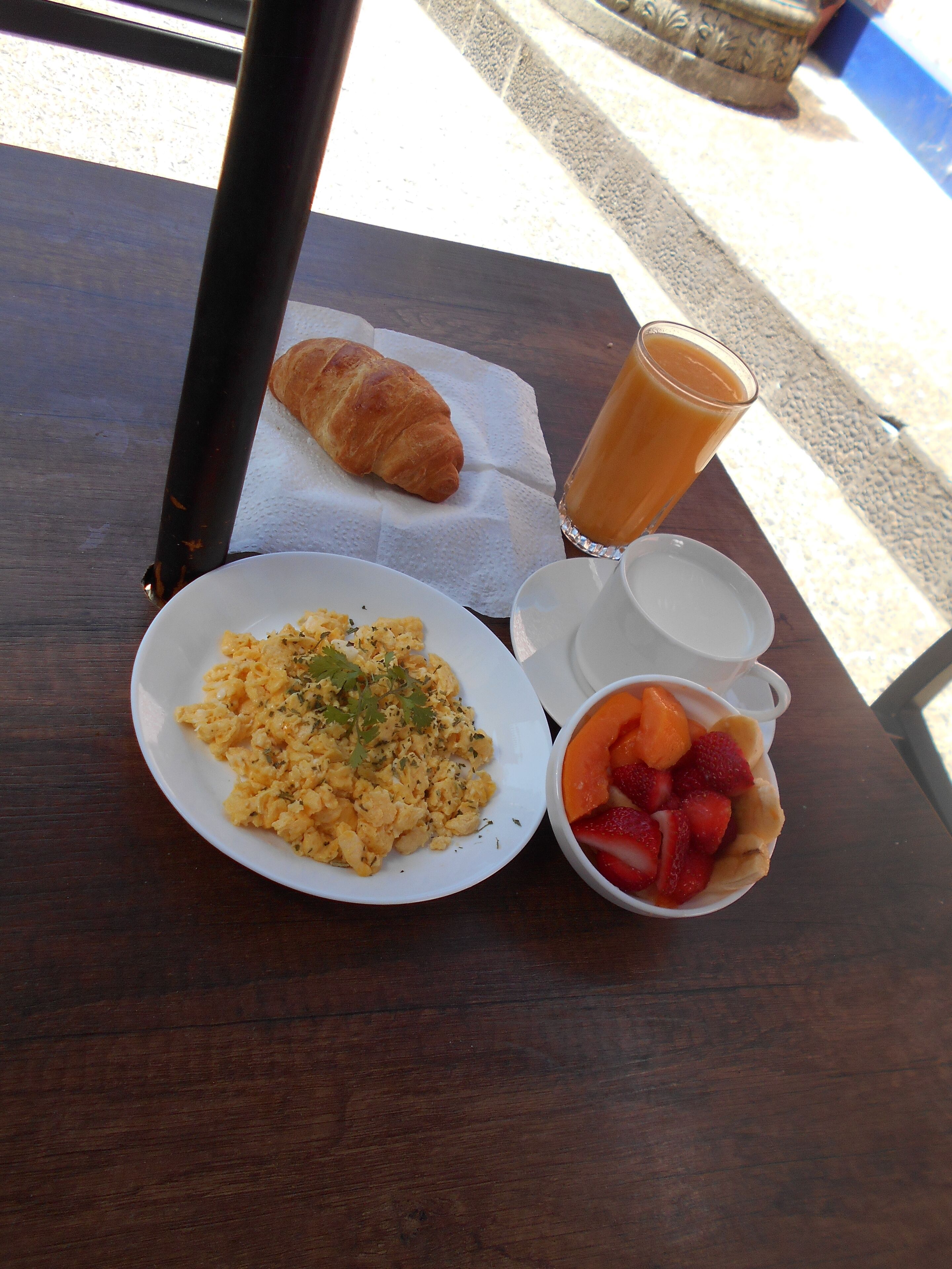 A breakfast table with scrambled eggs, a croissant, a glass of orange juice, and a bowl of fruit.