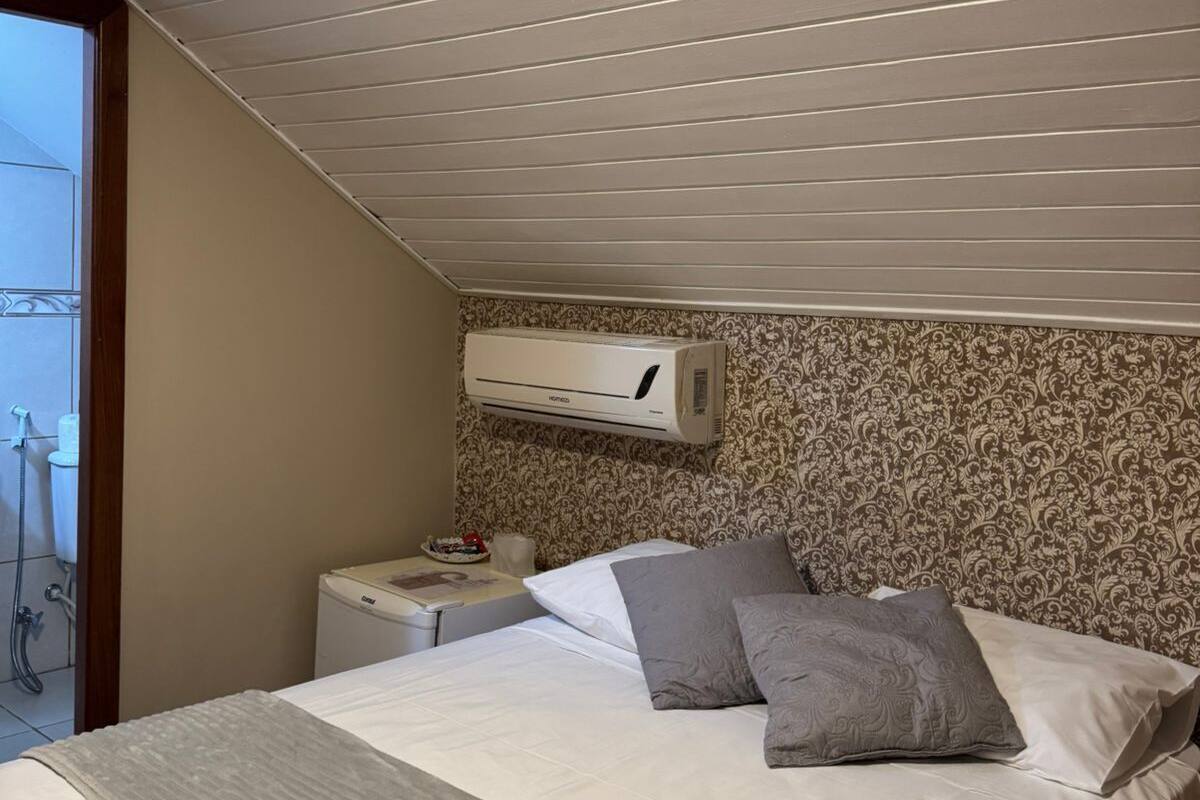 Une chambre avec un plafond en pente, un lit avec une literie blanche et grise, une table de chevet avec une lampe et un papier peint à motifs.