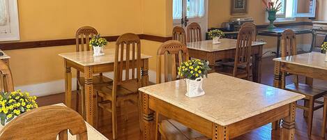 Uma sala de jantar com mesas e cadeiras de madeira, uma cozinha com fogão e pia, e uma janela com cortinas.