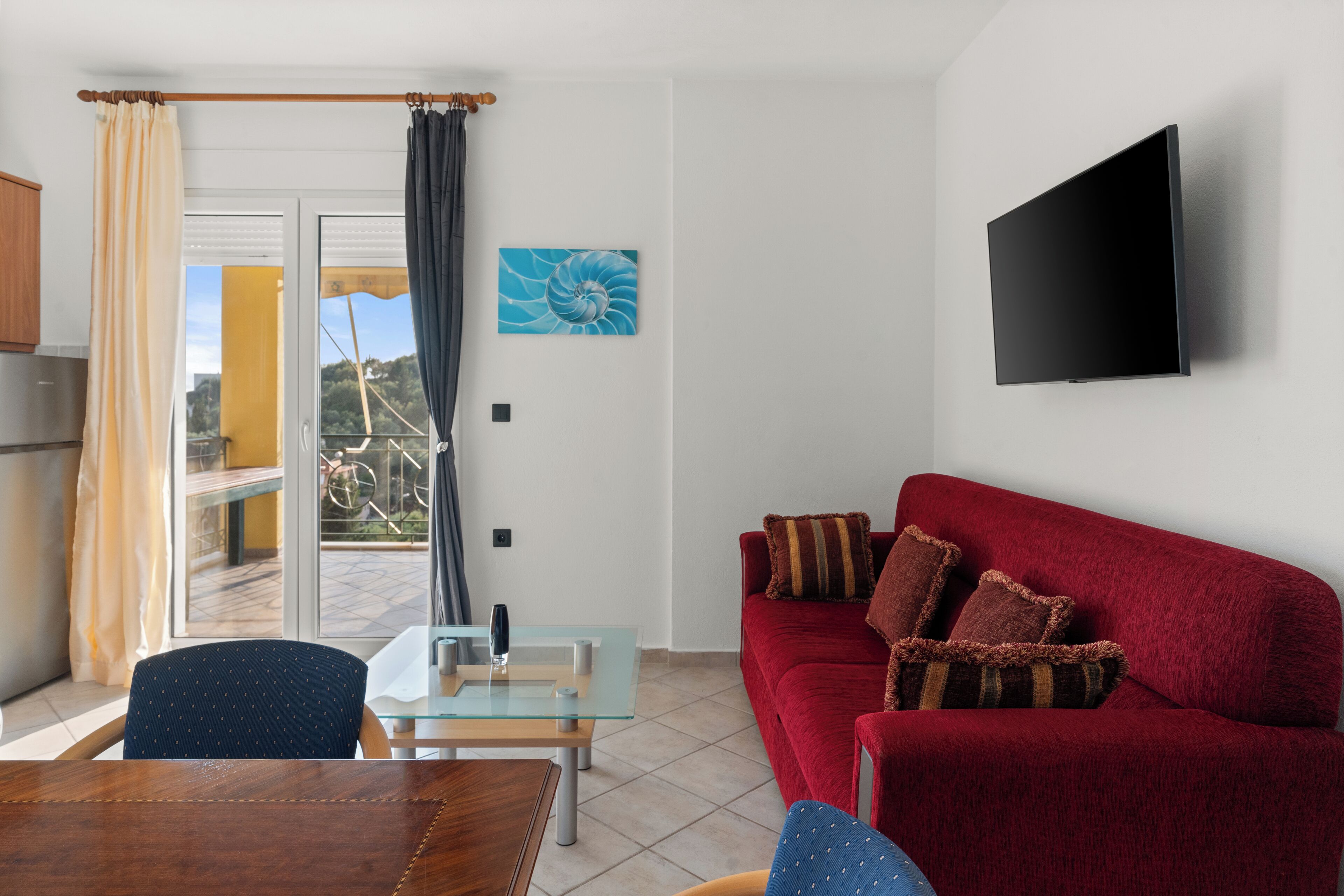 A living room with a red sofa, a glass coffee table, a wooden dining table, and a TV mounted on the wall.