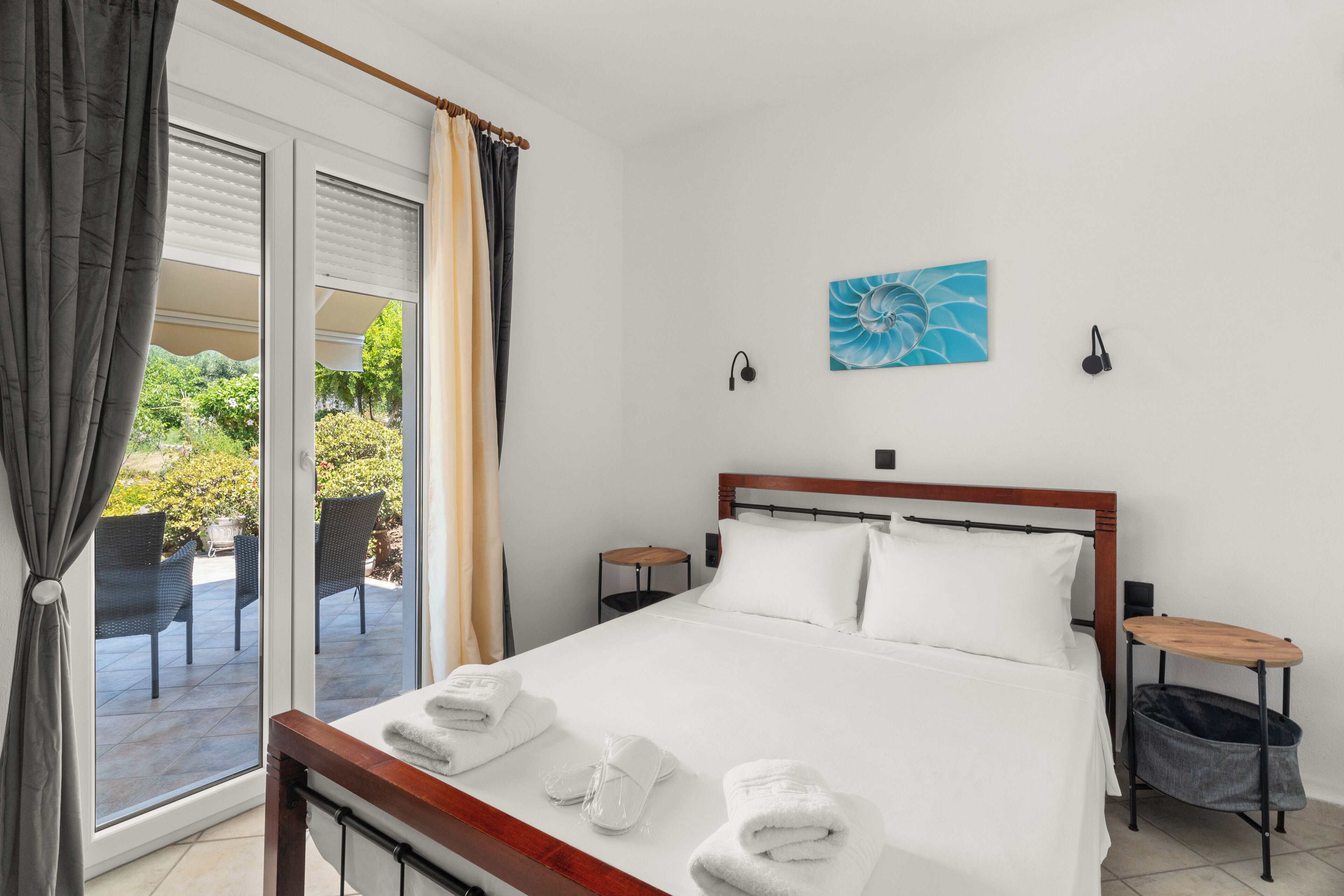 A hotel room with a bed, a nightstand, a small table, a trash bin, and a view of the outdoor patio.