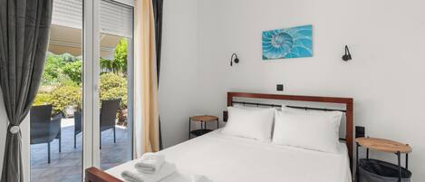 A hotel room with a bed, a nightstand, a small table, a trash bin, and a view of the outdoor patio.