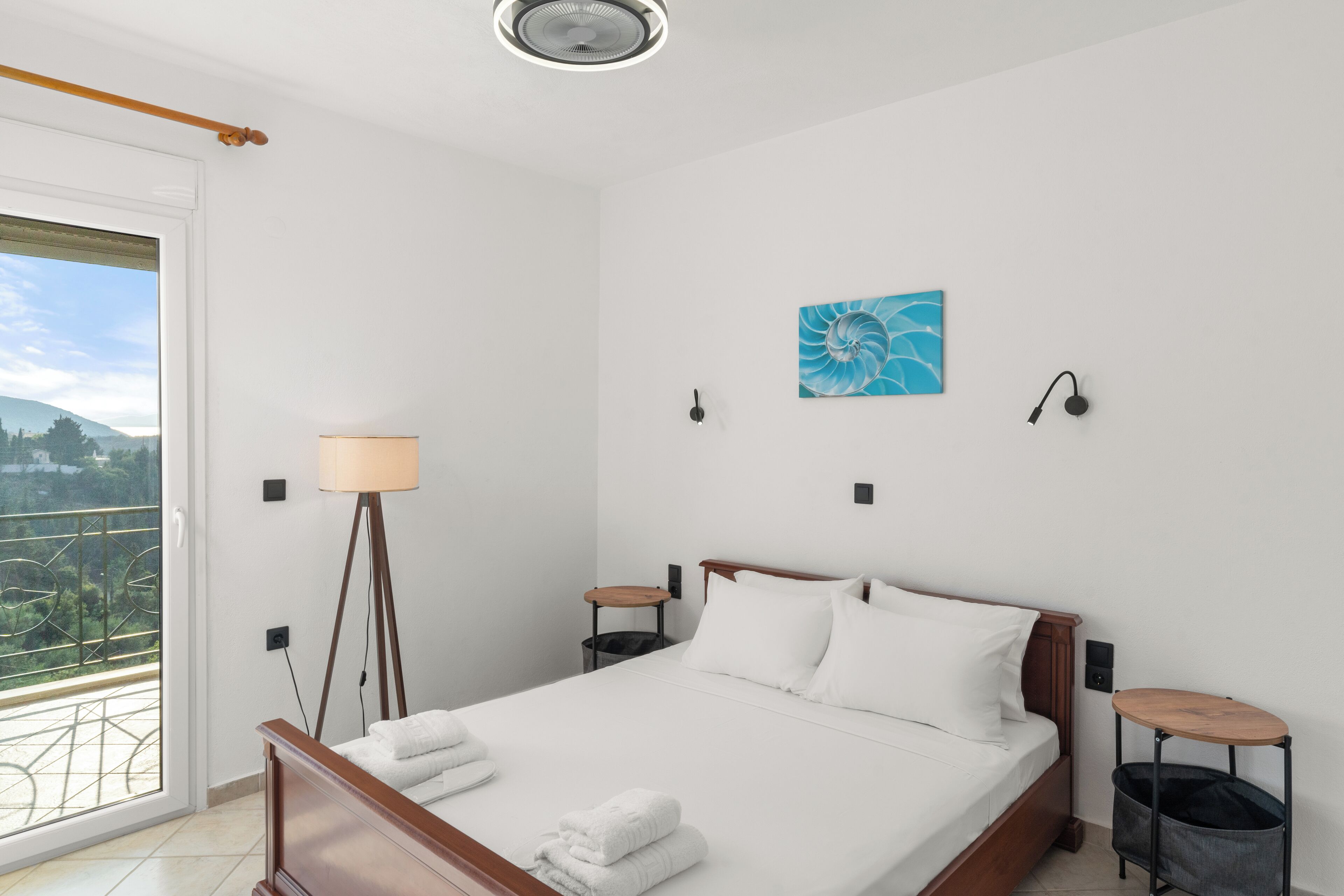 Un dormitorio con cama, mesita de noche, lámpara de pie, luz en la pared, un cuadro y vista al exterior a través de una ventana.