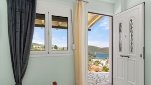 A bedroom with a bed, a window with curtains, a door leading to a balcony, and a view of the sea and hills. - The Dandy Horizon (Igoumenitsa)