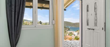Un dormitorio con cama, ventana con cortinas, puerta que da a un balcón y vistas al mar y las colinas.