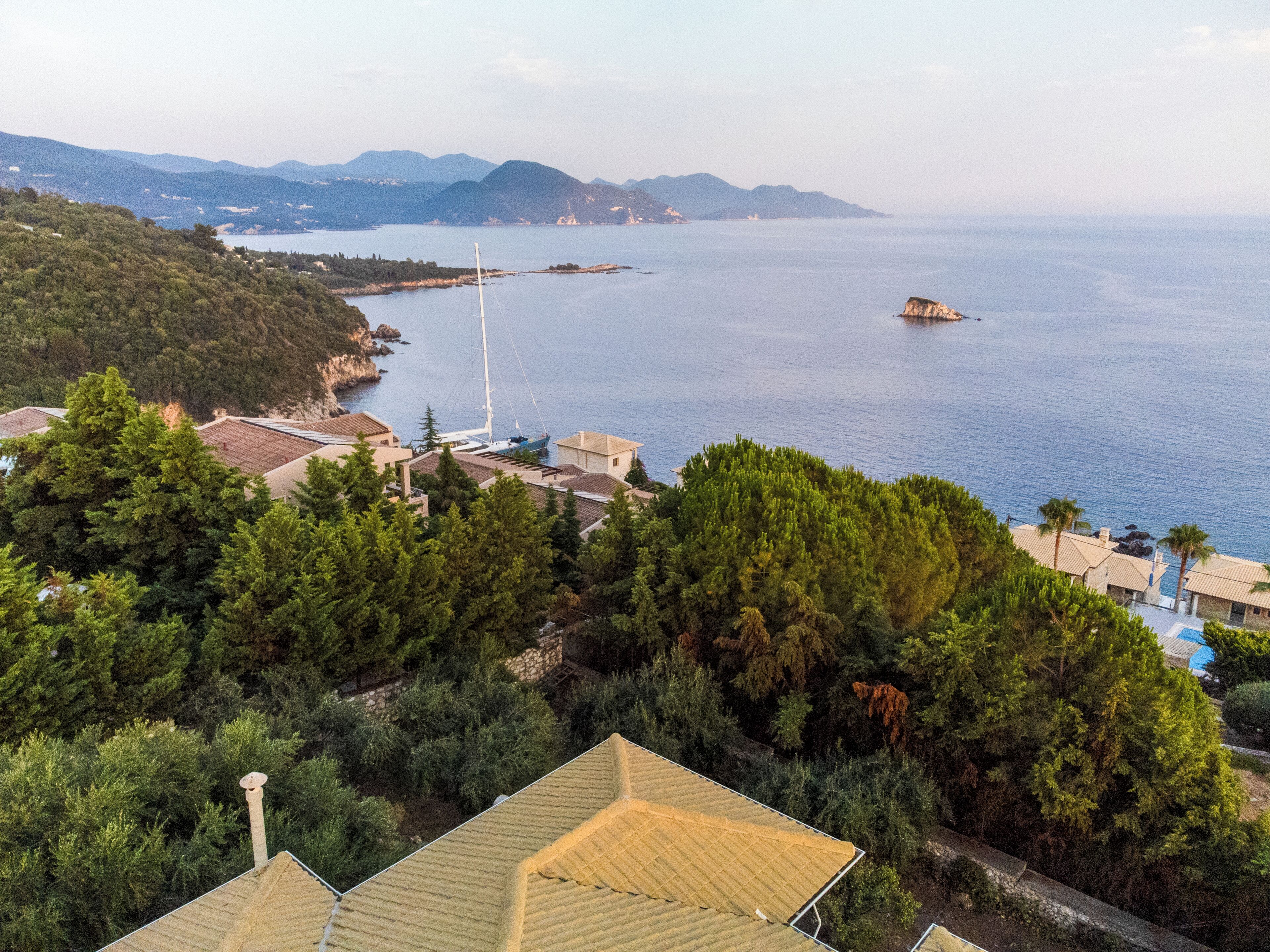 A coastal property with a view of the sea, a sailboat, and a mountainous backdrop.