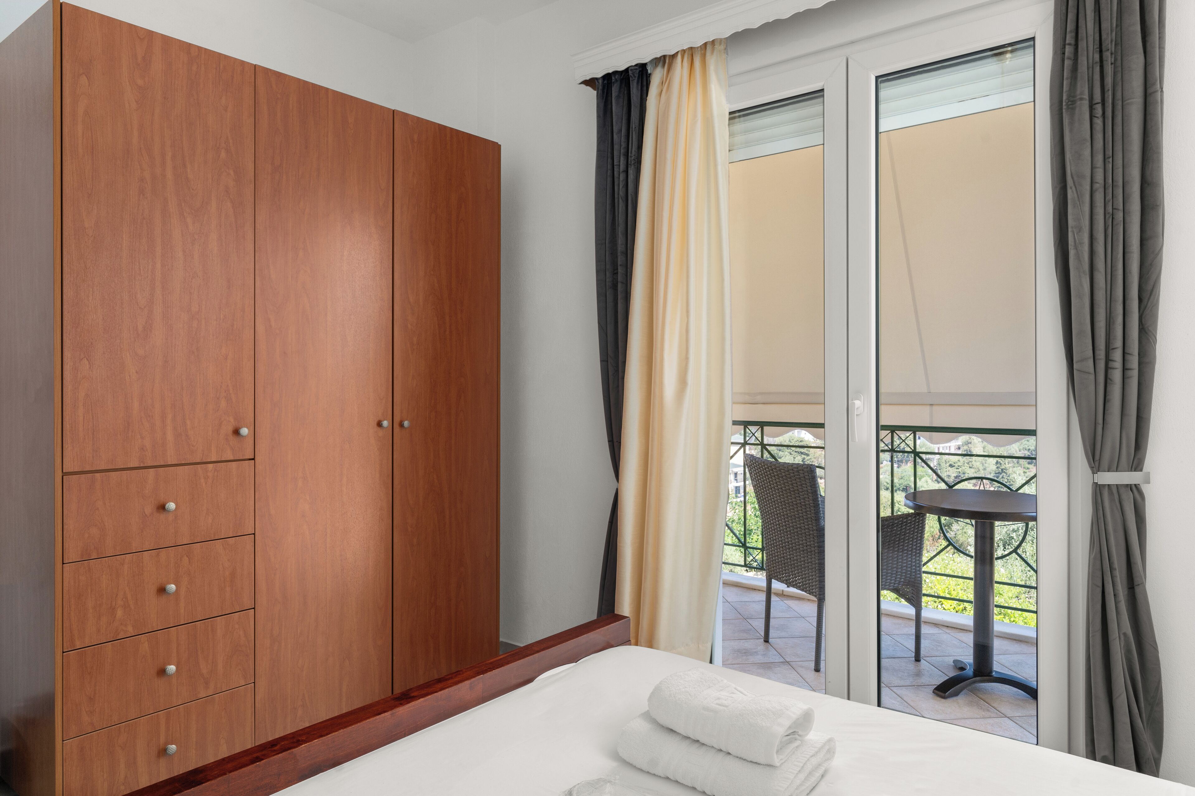 Un dormitorio con un armario de madera, una cama con ropa de cama blanca y un balcón con mesa y sillas.