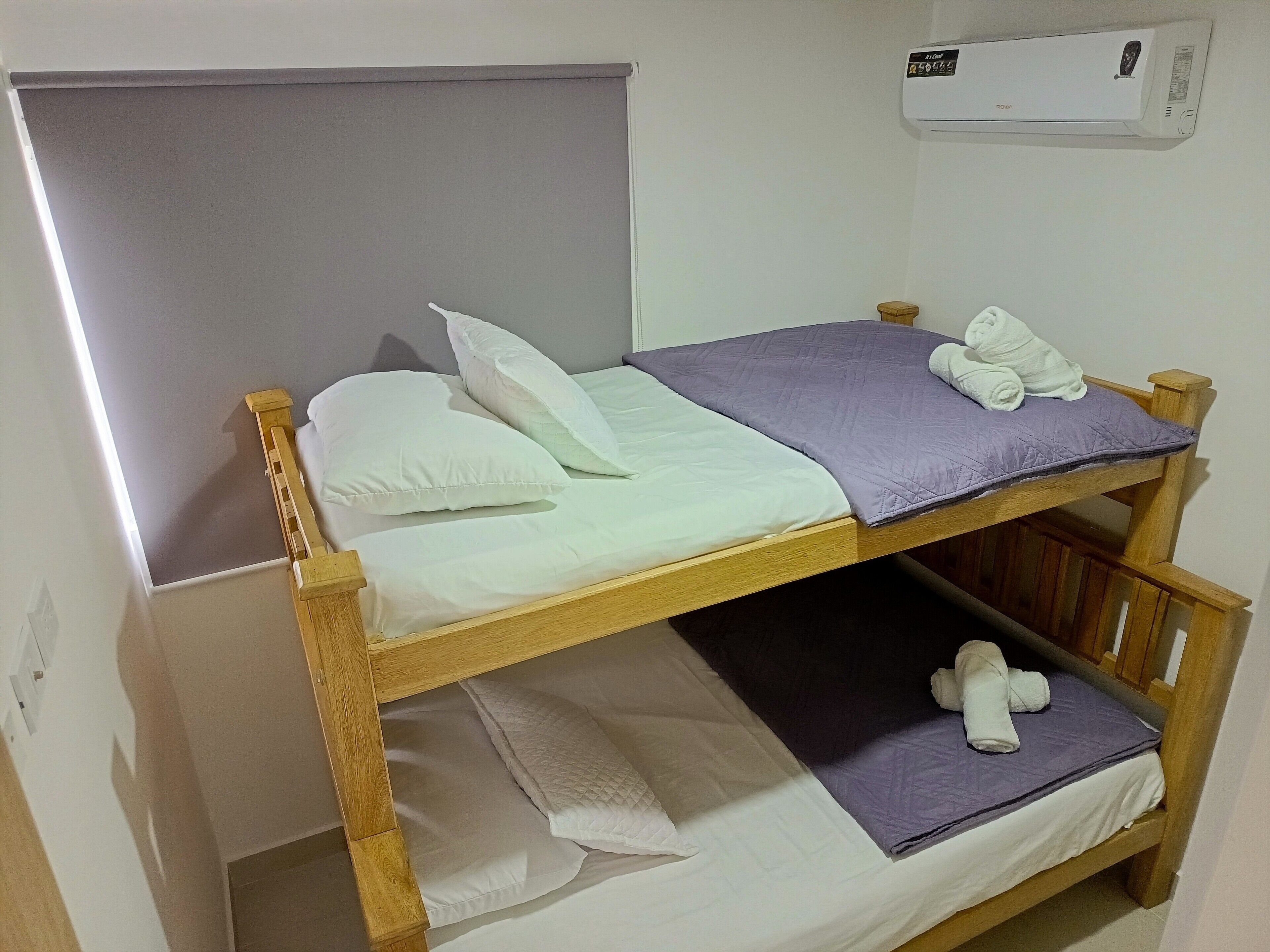 Een stapelbed met wit en paars beddengoed, een houten hoofdbord en een aan de muur bevestigde airconditioner.