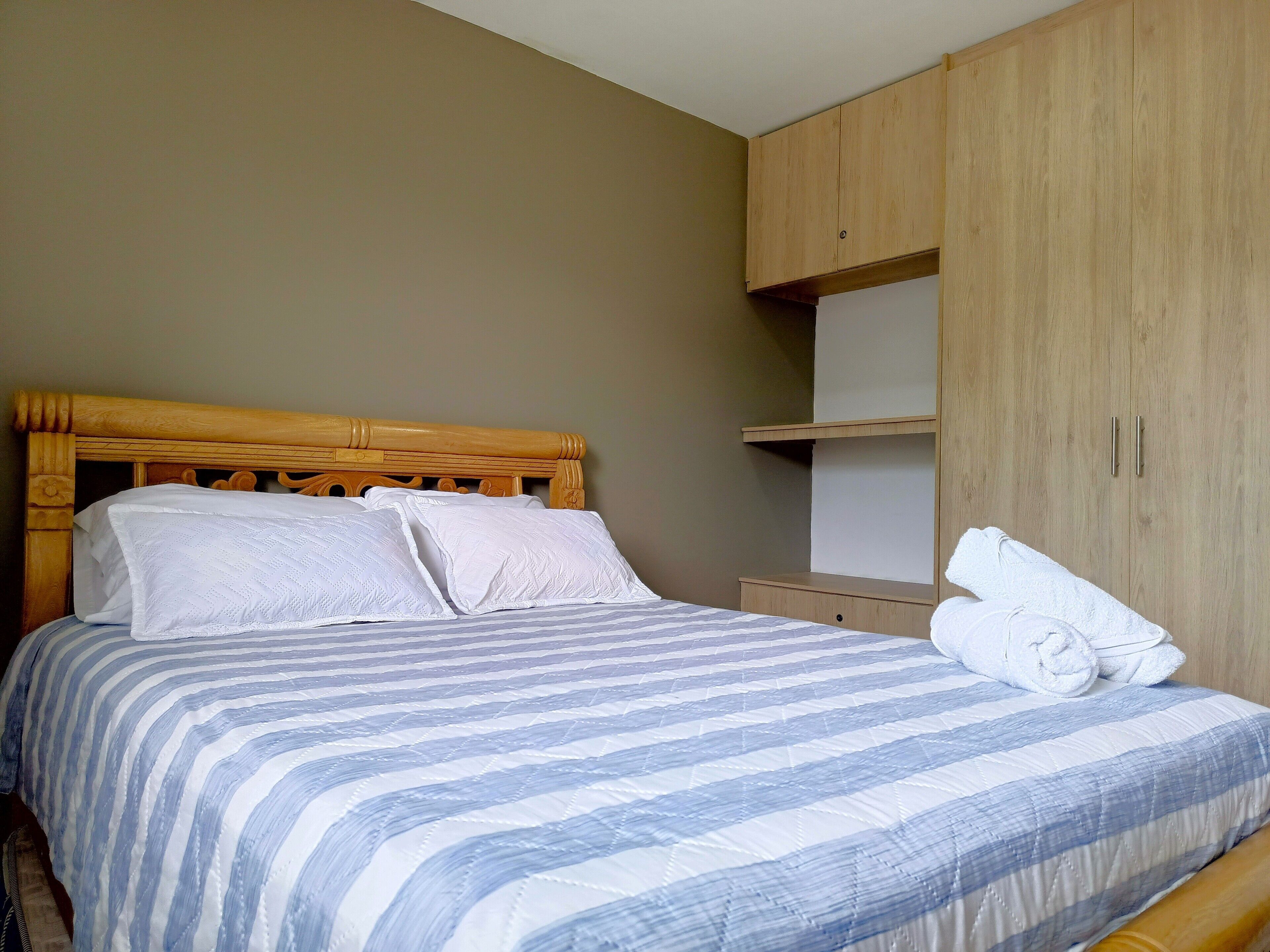 A neatly made bed with a striped bedspread, a wooden headboard, and a wooden wardrobe with a shelf above.