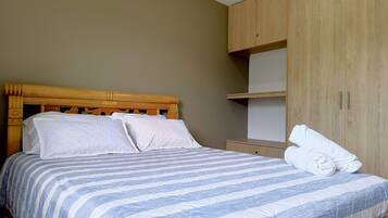 A neatly made bed with a striped bedspread, a wooden headboard, and a wooden wardrobe with a shelf above.