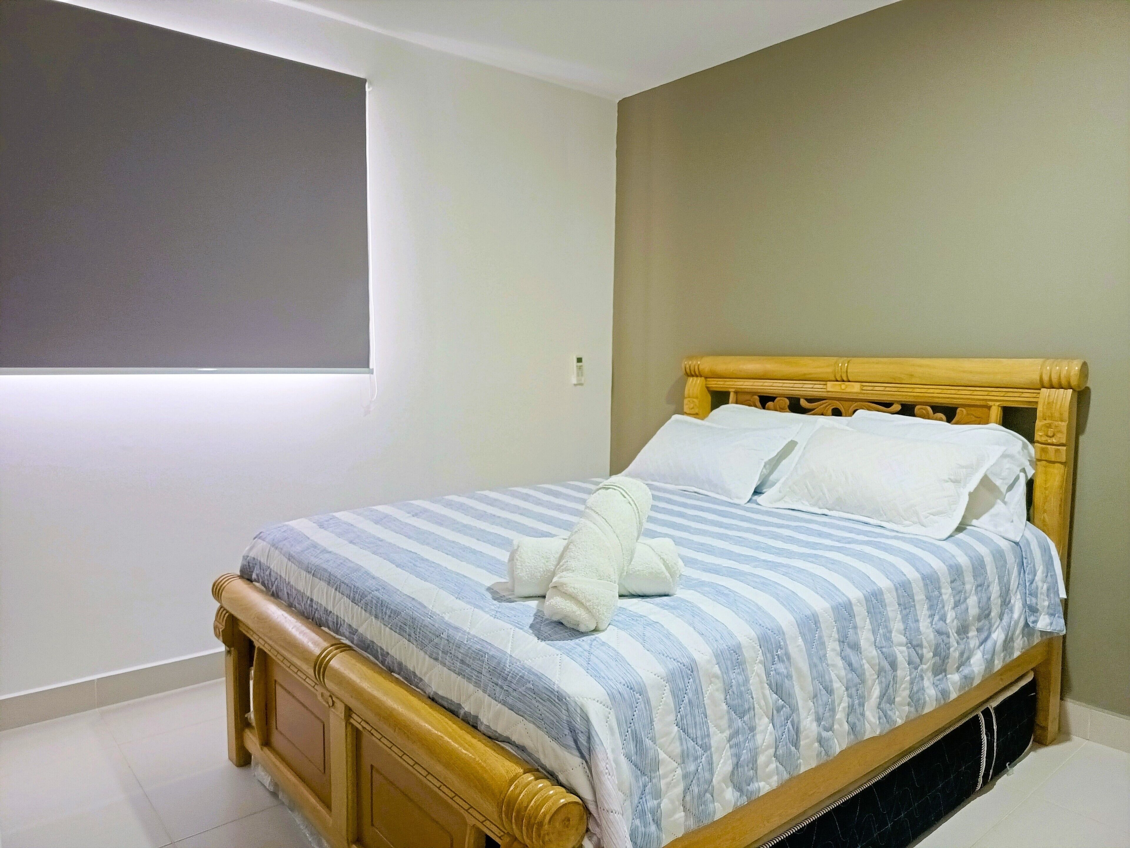 A neatly made bed with a striped bedspread and pillows, a wooden headboard, and a window with blinds.