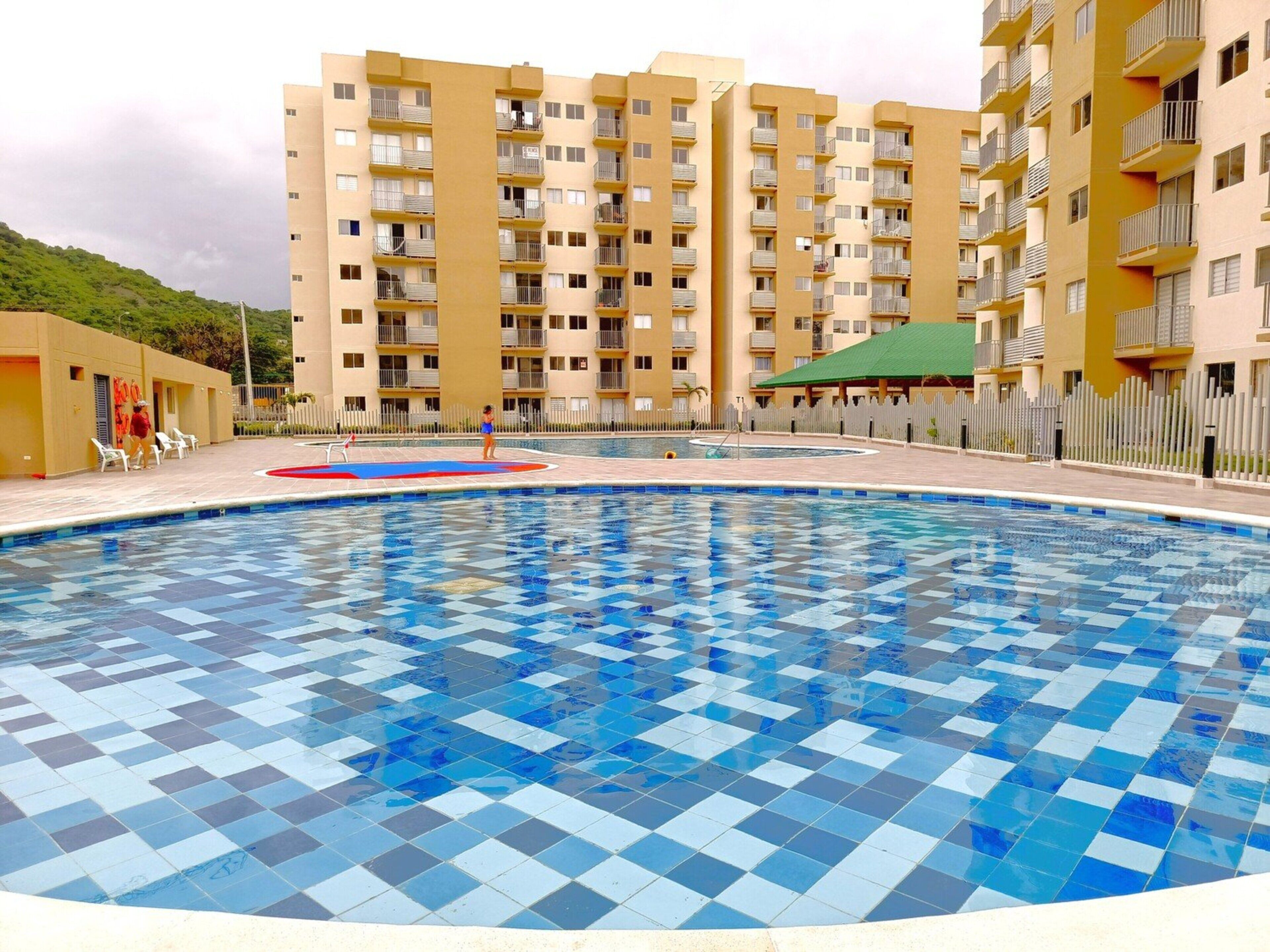 A swimming pool with blue checkered tiles, surrounded by a fence and a building with multiple balconies.