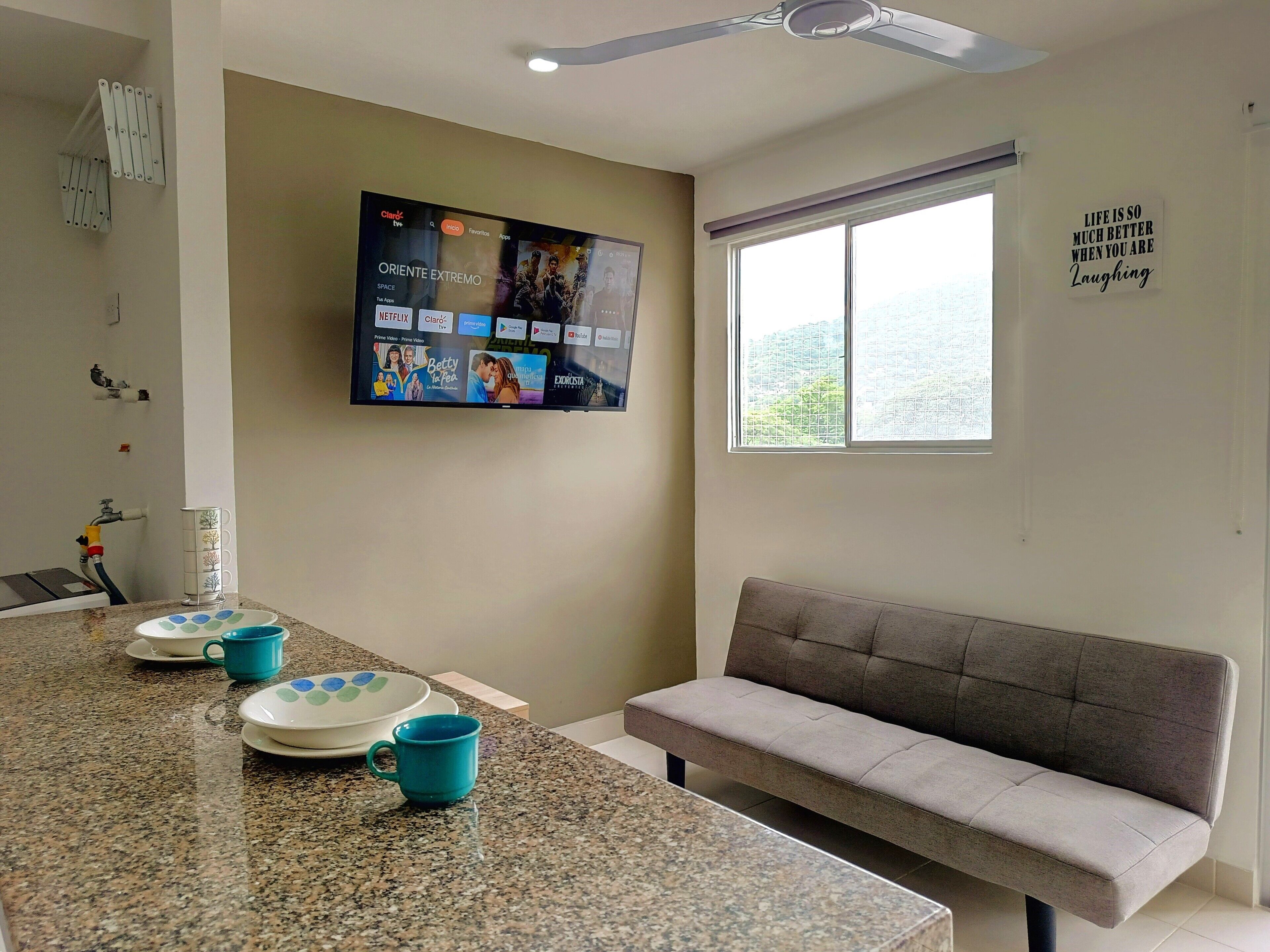 A modern living room with a flat-screen TV mounted on the wall, a grey sofa, a window with a view, and a kitchen area with granite countertops and blue mugs.