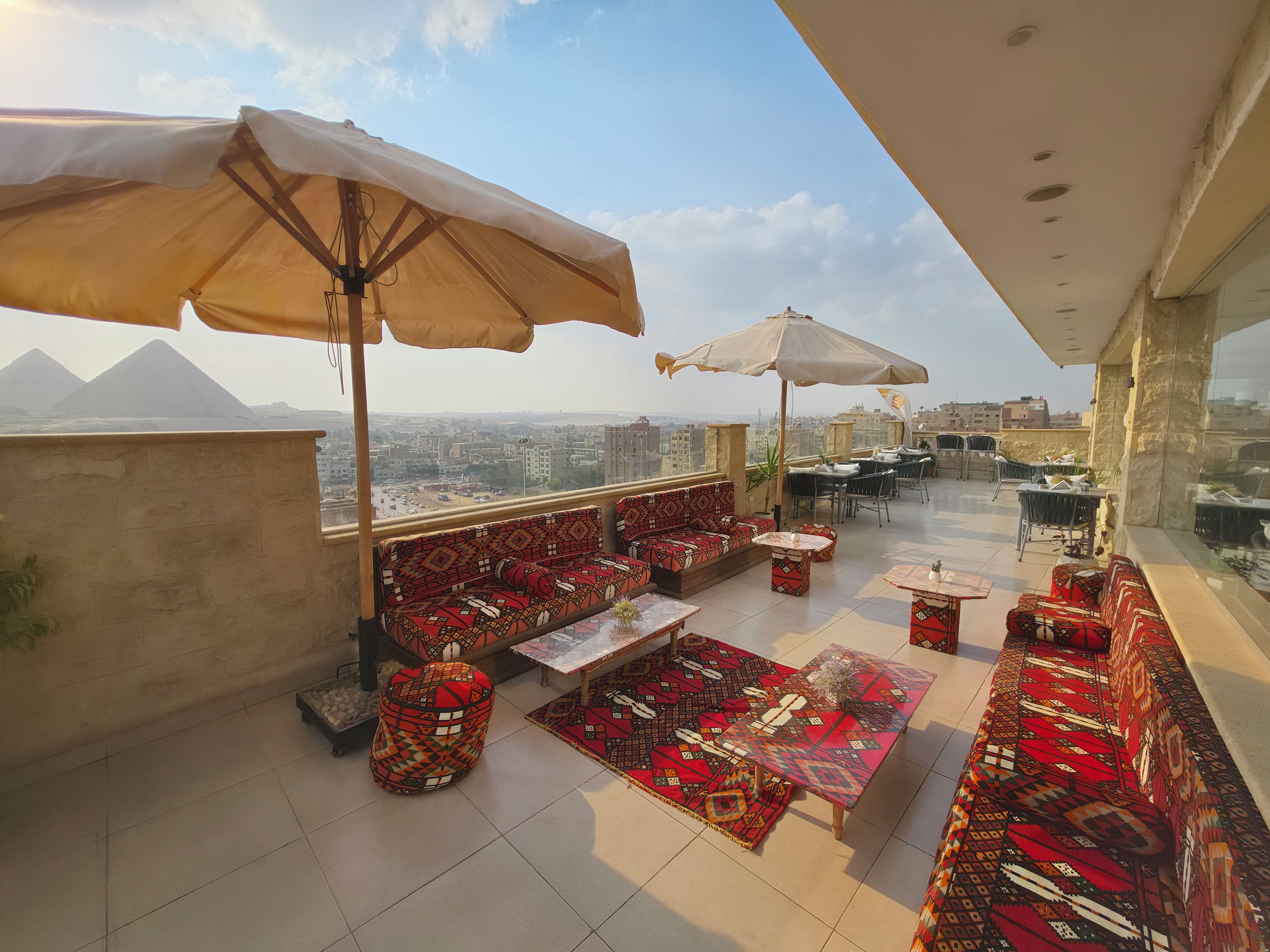 An outdoor terrace with seating, tables, and umbrellas, overlooking a cityscape and distant pyramids.
