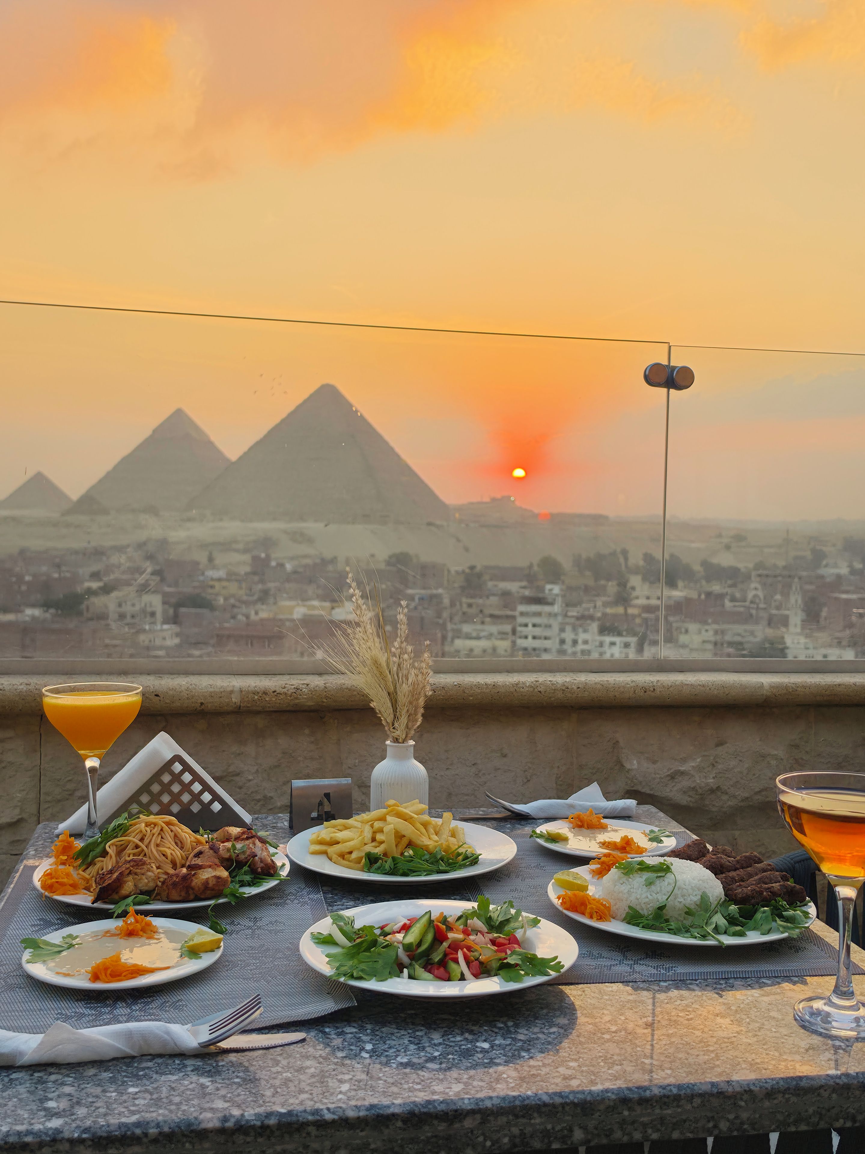 Una mesa con diversos platos y bebidas, con vista al atardecer sobre pirámides.