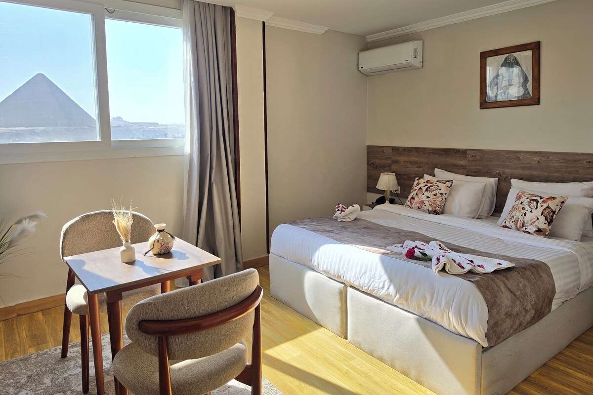 Ein Hotelzimmer mit Bett, Tisch mit Stühlen, Fenster mit Blick auf eine Pyramide und einer Klimaanlage.