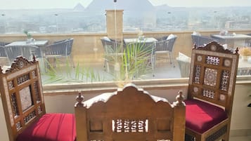 Un balcón con dos sillas de madera decorada, cojines rojos, una planta en maceta y vistas a pirámides a lo lejos.