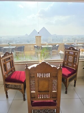 A balcony with two ornate wooden chairs with red cushions, a potted plant, and a view of pyramids in the distance.