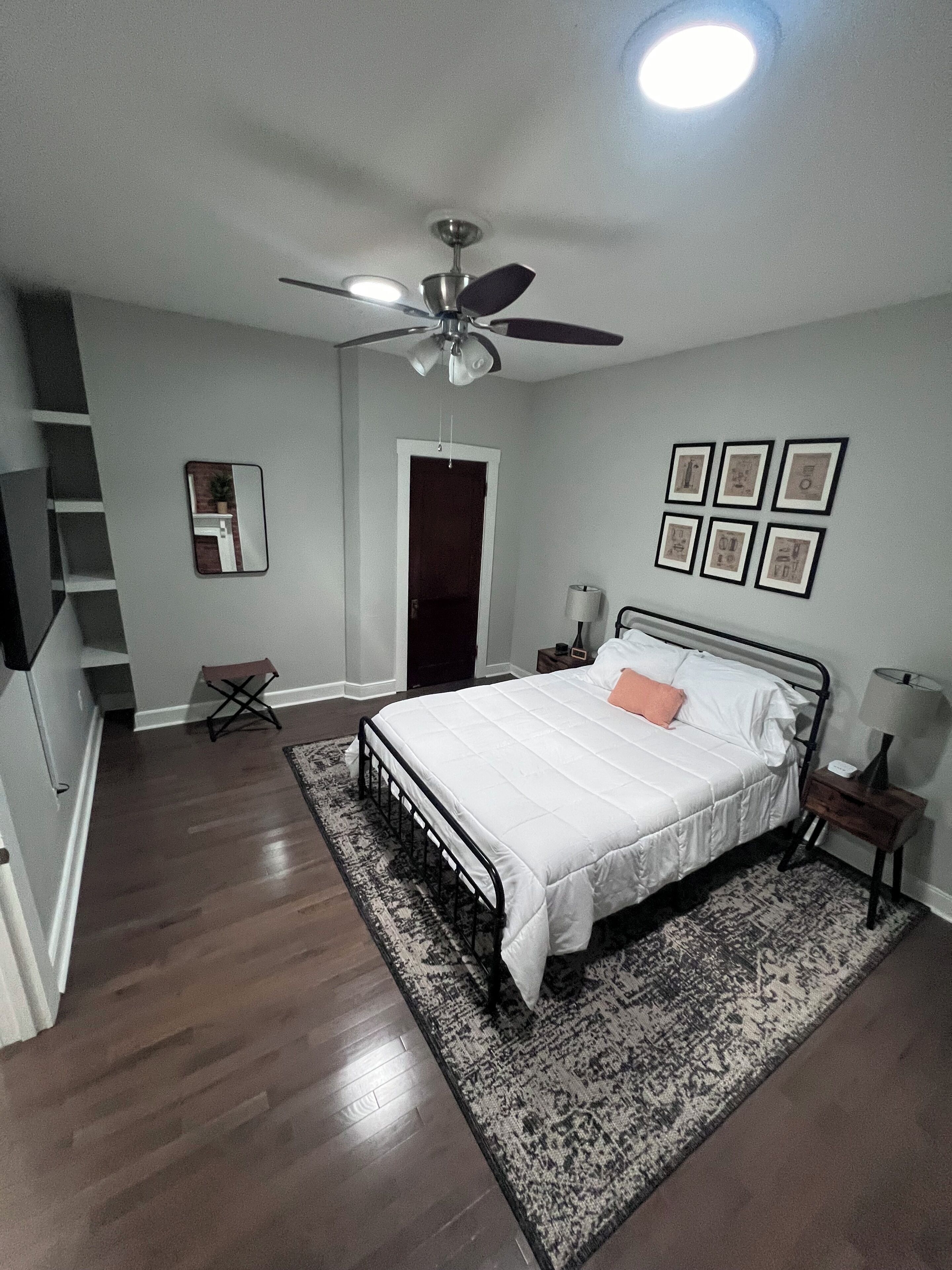 Ein Schlafzimmer mit einem Bett, Deckenventilator, Nachttisch, Lampe, Spiegel und gerahmten Bildern an der Wand.
